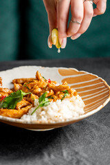 A close-up of a hand squeezing fresh lime juice over a plate of chicken served with rice and garnished with herbs. The dish is vibrant and flavorful on a textured surface.