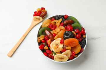 Mix of different dried fruits in bowl and spoon on white marble table, flat lay