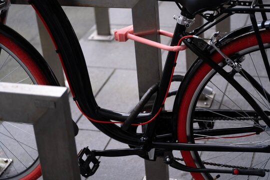 A partial view of a bicycle secured to a metal stand with a pink U-lock. The focus is on the frame, lock, and part of the wheel