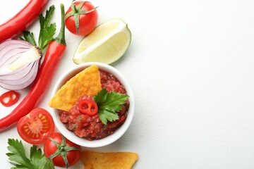 Spicy salsa sauce in bowl, nachos and ingredients on white table, flat lay. Space for text