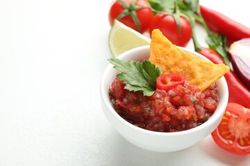 Spicy salsa sauce in bowl, nacho chip and ingredients on white table, closeup. Space for text