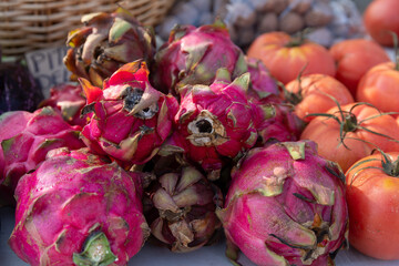 Bright pink dragon fruits with spiky green-tipped skin piled on a market stand, with tomatoes and...