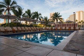 Serene pool with lounge chairs, straw umbrellas, and striped cabanas, surrounded by palm trees. High rise buildings visible in the background.