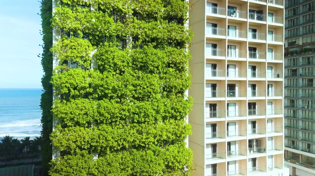 Modern building stands tall with its facade entirely covered in vibrant green vegetation, offering a striking contrast to the nearby ocean and other buildings