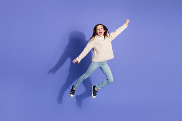 Playful young girl jumping against a purple background in casual sweater and jeans showcasing energetic fun