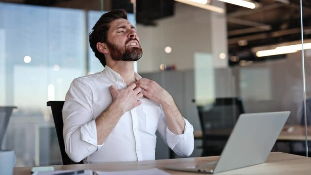 Frustrated businessman with a beard scratches and rubs his neck and shoulder while sitting at his desk in a modern office. He then throws his hands up in exasperation and continues typing on his