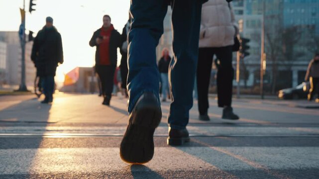 Back view following male feet steps walking pedestrian crossing street on sunny autumn day. Man wearing leather boots goes crossroad. Low angle people legs stepping backlit warm sunlight