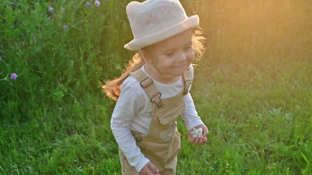 Cute toddler girl in beige overall and hat dancing joyfully in a sunlit field of tall grass during a warm and soft summer sunset