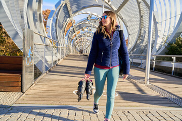 Young sporty woman walking on modern bridge carrying inline skates