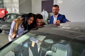 Smiling couple enthusiastically examines a new car with a salesman at a dealership, showcasing...