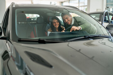 Couple sits inside a sleek new car at a dealership, enthusiastically exploring its features. The...