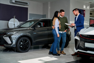 Young couple discusses options with a car salesman inside a modern showroom. They stand near two...