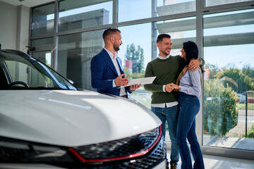 Joyful couple stands with a salesman in a car dealership, holding documents and smiling,...