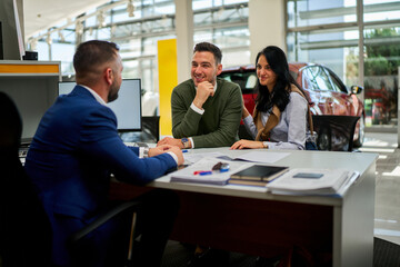 Enthusiastic young couple discussing car purchase options with a professional salesman at a modern dealership. Bright showroom environment with a sleek vehicle in the background