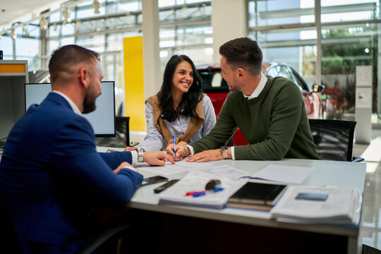Business professionals engaging in a discussion around a car purchase agreement at a dealership. Smiling woman signing documents while two men provide assistance and advice