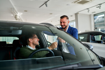 Smiling car salesman in a dealership hands over keys to a satisfied customer sitting in a new vehicle. The interaction highlights a successful car purchase and customer service experience