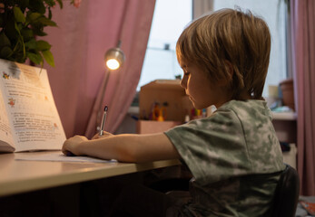 A 6-year-old boy sits at the table and carefully writes his homework. Schoolboy doing homework in...