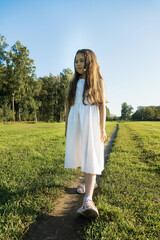 A young girl with long wavy brown hair stands in a sun-drenched green field in a white dress. Sunlight illuminates her hair.