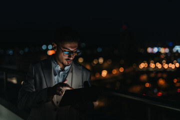 A focused business professional uses a tablet while standing outdoors at night. The urban setting...