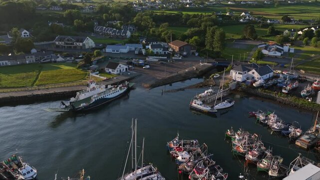 Lough Foyle Ferry, County Donegal, Ireland, June 2023. Drone orbits counter clockwise above the harbour at Greencastle with fishing boats and yachts docked along the piers at golden hour.