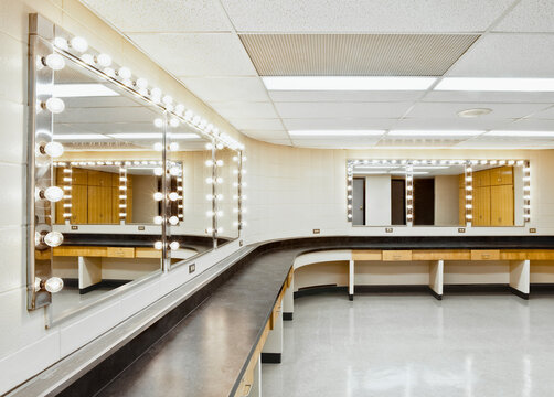 A theatre dressing room with a long worktop and seats and big mirrors with bulb lighting around the edge.