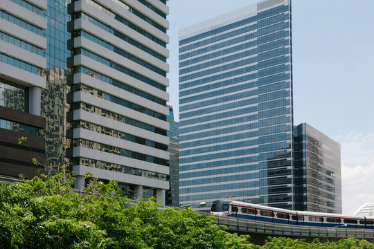 A mass transit train on an elevated track passes modern glass buildings in the central business district of Bangkok at treetop level.