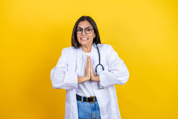 Young brunette doctor woman wearing stethoscope standing over isolated yellow background begging and praying with hands together with hope expression on face very emotional and worried
