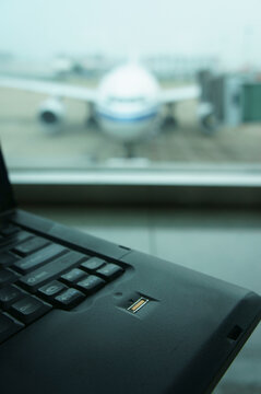 A computer laptop on a surface,in front of a window. Airport apron and aircraft seen through the window.