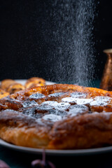 Banana Tarte Tatin is sprinkled with powdered sugar on a white plate on a dark background. The powder is poured onto the cake. Vertical Photo