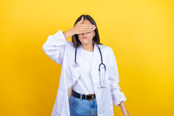 Young brunette doctor woman wearing stethoscope standing over isolated yellow background serious and covering her eyes with her hand