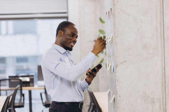 Young african american manager taking notes on colorful sticky notes attached to a wall in a modern office, using a pen and consulting his smartphone