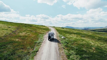 Drone follows 4x4 camper van drive on a gravel mountain road on sunny day with blue skies and clouds. Road trip adventure in overlanding van with solar setup on roof rack. Overland travel 