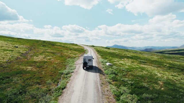 Drone follows 4x4 camper van drive on a gravel mountain road on sunny day with blue skies and clouds. Road trip adventure in overlanding van with solar setup on roof rack. Overland travel 