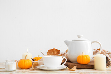 Tea pot, cup of hot drink, pumpkins, burning candles and autumn leaves on table near white wall