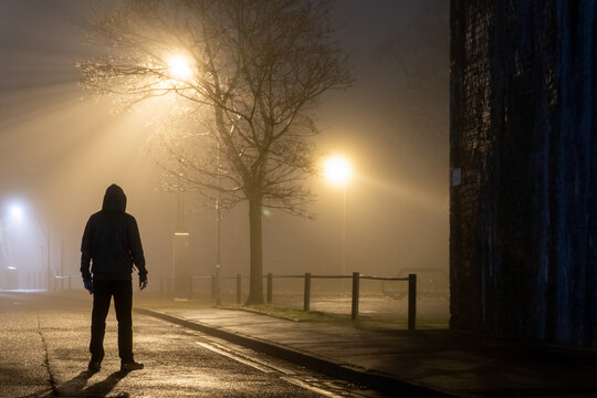 A sinister hooded figure, standing on a empty city road. On a mysterious foggy night. Silhouetted by street lights