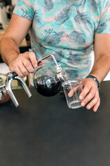 Close up of unrecognizable barista expertly pours brewed coffee from Japanese siphon coffee maker into a glass, showcasing the art of specialty coffee preparation. Copy space at the bottom of image.