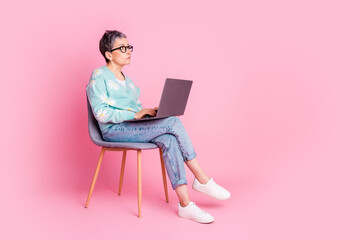 Stylish elderly woman with grey hair using laptop seated against a pink background wearing casual...