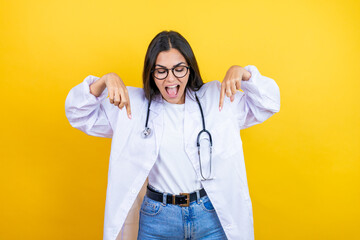 Young brunette doctor woman wearing stethoscope standing over isolated yellow background surprised, looking down and pointing down with fingers and raised arms