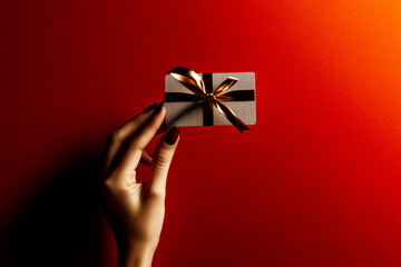 A woman's hand with red nails holds a luxurious gift card wrapped with a gold ribbon against a rich red background, symbolizing elegance, exclusivity, and premium gifting for special occasions .