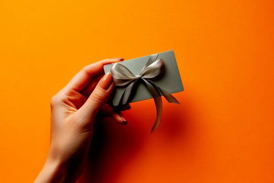 A woman's hand with elegant nude nails holds a stylish gift card wrapped with a delicate silver ribbon against a warm orange background, symbolizing premium gifting, elegance.
