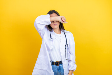 Young brunette doctor woman wearing stethoscope standing over isolated yellow background covering eyes with arm smiling cheerful and funny. Blind concept.