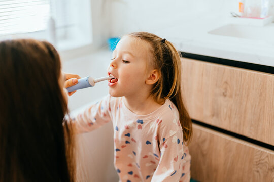little girl, 4 years old, sits near bathroom sink, mother carefully helps brush her teeth with electric toothbrush. Intimate moment of daily care, highlighting the importance of oral hygiene for kids.