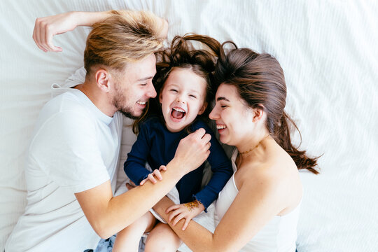 Happy family of three lying on bed, laughing and enjoying playful moment. Father and mother smile, little daughter giggles, feeling safe and loved in embrace. Family connection concept.