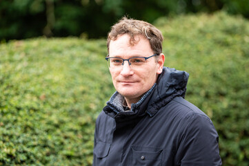 Portrait of a 45 yo white male wearing a dark winter jacket against a  green nature background, Koekelberg, Brussels, Belgium