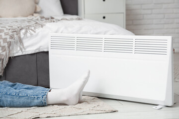 Young woman warming her feet near electric convector heater in bedroom