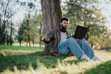 A man appears frustrated while using his laptop outdoors, sitting under a tree in a park setting. Sunny day and nature background emphasize his emotions, likely due to technical or connectivity issues