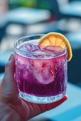 A person is holding a glass of purple drink with a slice of lemon on top. The drink appears to be a purple cocktail, possibly a purple martini or a purple smoothie