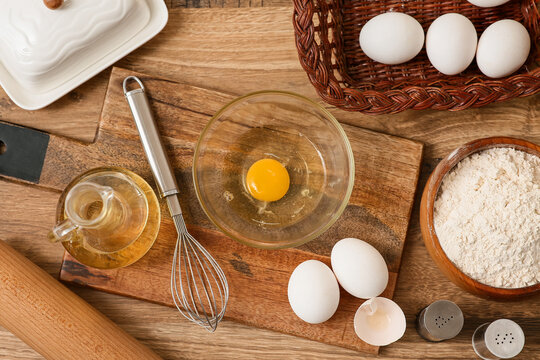 Bowl of flour with whisk, eggs and jug of oil for preparing dough on wooden background
