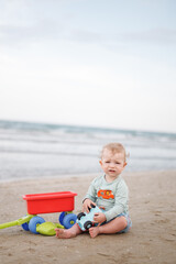 one year-old child, boy, sitting at the sea shore at sunny summer day and playing a bucket of water. Baby at the sea background. Naturist