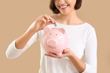 Young woman putting coin into piggy bank on beige background, closeup